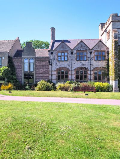 Stone school range with triple gables, garden bench and sunlit lawn.
